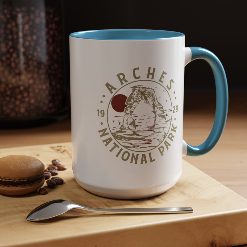 White ceramic mug with a contrasting handle and interior, featuring a design of Arches National Park with Delicate Arch and a sun graphic.