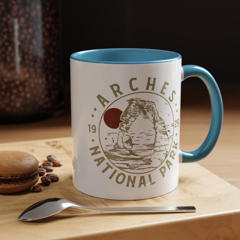 White ceramic mug with a contrasting handle and interior, featuring a design of Arches National Park with Delicate Arch and a sun graphic.