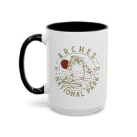 White ceramic mug with a contrasting handle and interior, featuring a design of Arches National Park with Delicate Arch and a sun graphic.